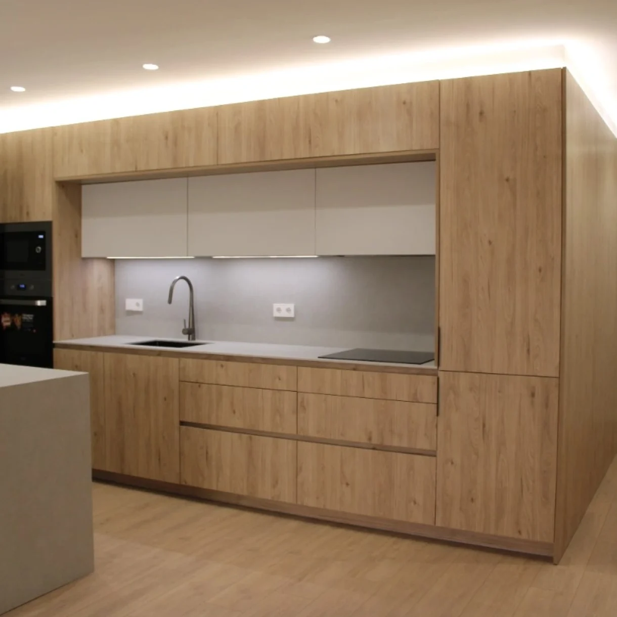 Cocina moderna en proyecto Sicília, Barcelona, con armarios de madera clara, encimera gris y electrodomésticos integrados.