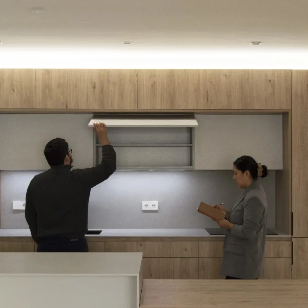 Pareja inspeccionando cocina moderna con gabinetes de madera clara y encimeras grises en proyecto Sicília