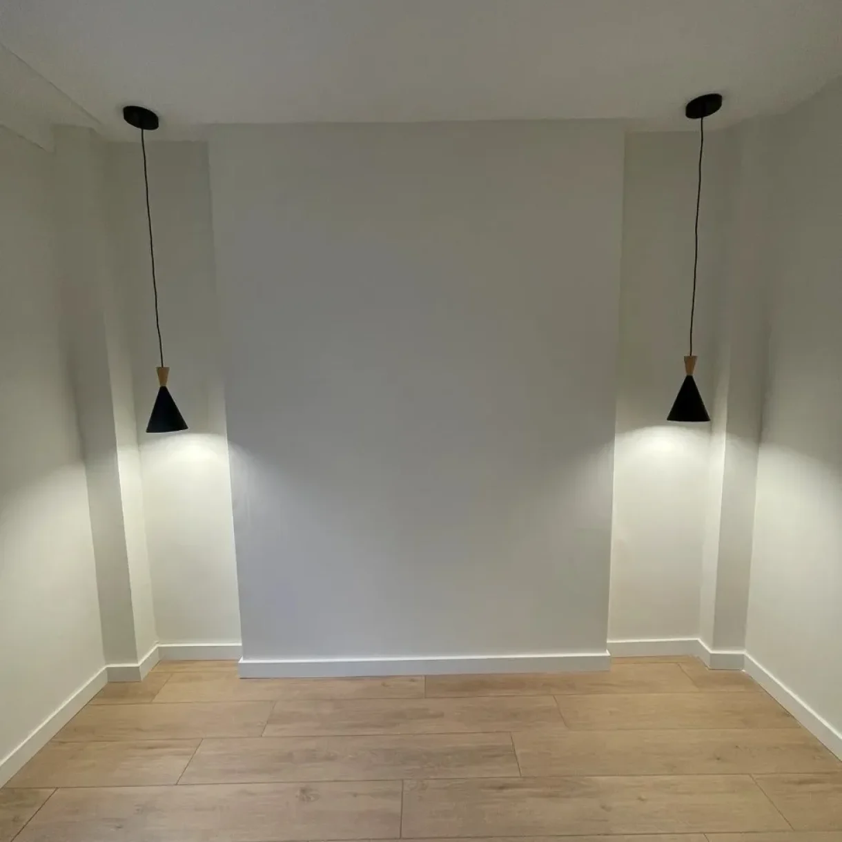 Habitación vacía con paredes blancas, suelo de madera clara y dos lámparas colgantes negras en proyecto Sicília, Barcelona