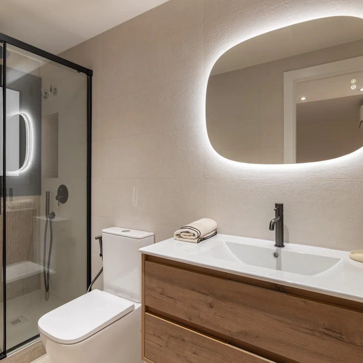 Baño moderno y funcional, diseñado con revestimientos en tonos neutros y muebles a medida, con un plato de ducha amplio y mampara de cristal, optimizando el espacio en la reforma integral de un piso en Sant Andreu.